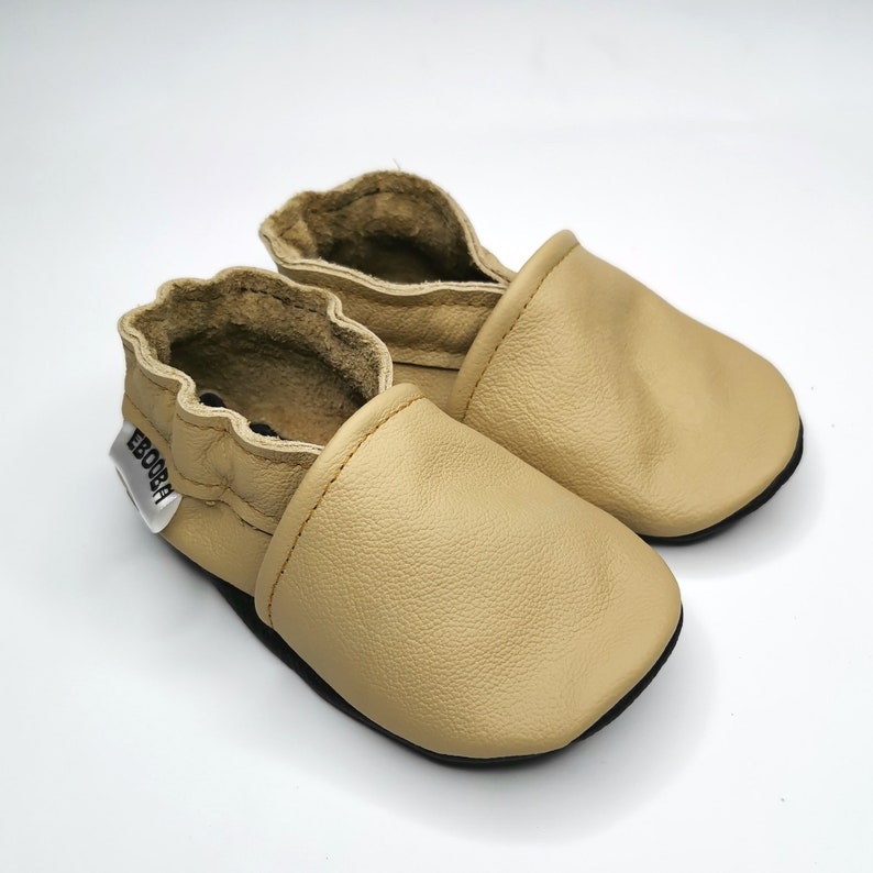 May include: A pair of beige leather baby shoes with a soft, flexible sole. The shoes have a simple design and are perfect for crawling and walking. The shoes are made by the brand "Ebooba".