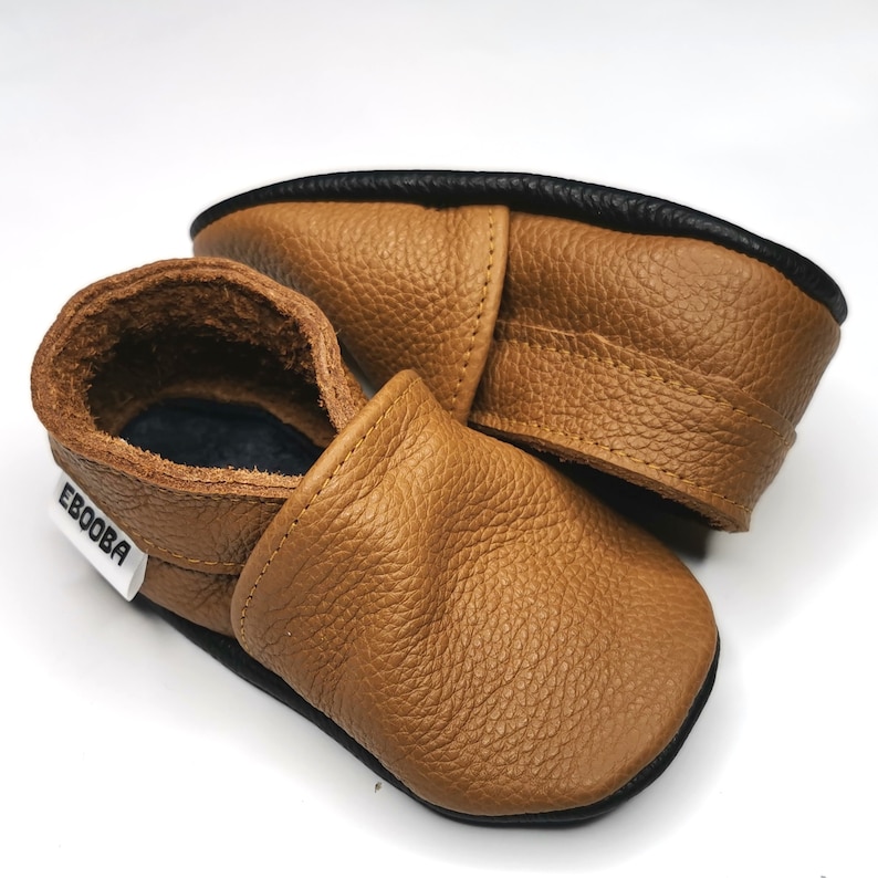 May include: A pair of brown leather baby shoes with a black trim. The shoes have a slip-on design and a label that says "Ebooba".