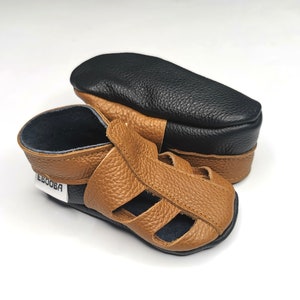 May include: Tan leather sandals with a black leather strap and the word "Ebooba" on the strap. The sandals have a cut-out design and are made of soft leather.