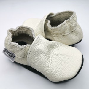 May include: A pair of white leather baby shoes with a black sole and the word "Ebooba" on the side.