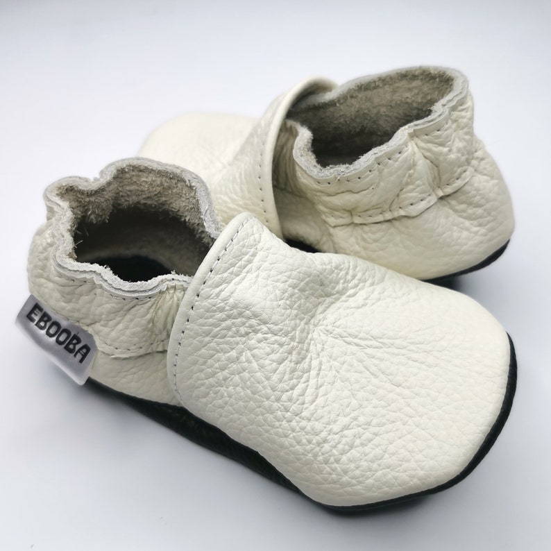 May include: A pair of white leather baby shoes with a black sole and the word "Ebooba" on the side.