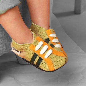 Puede incluir: Zapatillas de bebé en verde oliva claro con detalles en naranja y negro. Los zapatos tienen cordones blancos y la marca "EBOOBH" es visible en una etiqueta. Los zapatos están hechos de un material perforado.