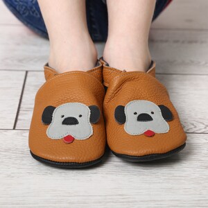 Peut inclure: Chaussons pour bébé en cuir marron avec un motif de visage de chien. Les chaussures ont un visage gris, des oreilles noires et une langue rouge. Les semelles sont noires.