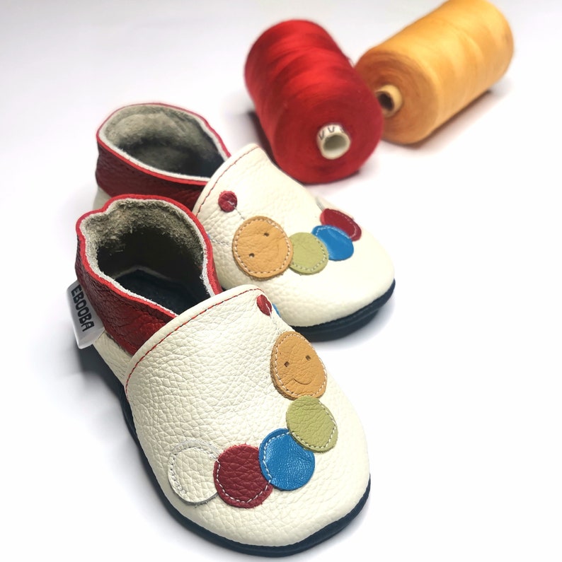 May include: White leather baby shoes with a red trim and colorful smiley face buttons. The shoes are on a white background with spools of red and orange thread.