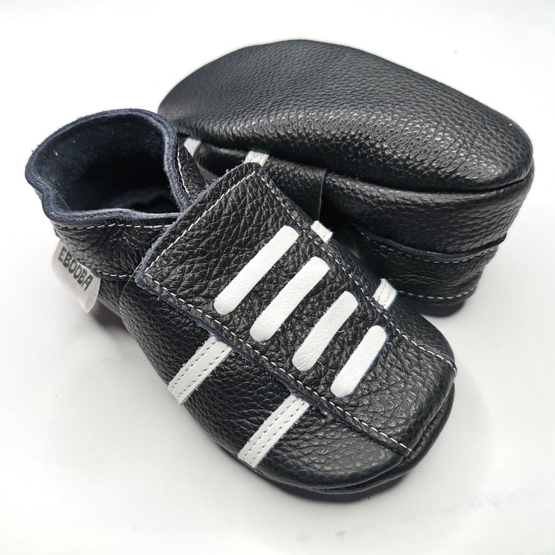 May include: Black leather baby shoes with white stripes and a velcro closure. The shoes have the word "Ebooba" on the side.