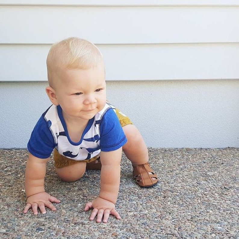 May include: A baby boy wearing a blue and white striped shirt with a brown and yellow patterned shorts, crawling on a gray surface. He is wearing brown sandals.