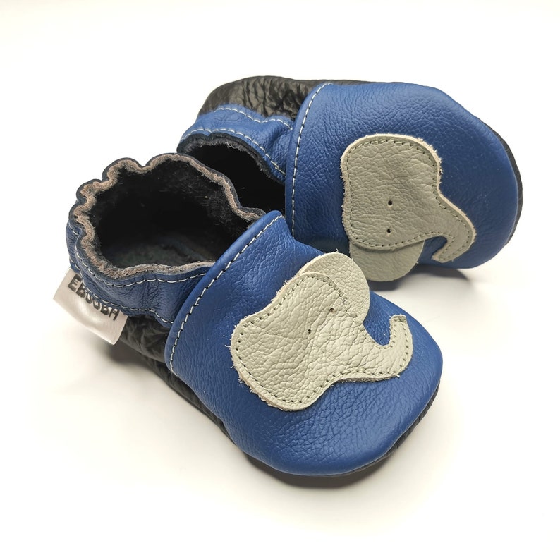 K&ouml;nnte beinhalten: Ein Paar blaue Lederschuhe f&uuml;r Babys mit einem grauen Elefanten-Design. Die Schuhe haben eine weiche, flexible Sohle und sind perfekt f&uuml;r kleine F&uuml;&szlig;e.