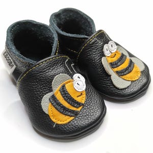 May include: Black leather baby shoes with a bee design. The bee is yellow and black with grey wings. The shoes have a yellow stitch around the edge and a black and grey lining.