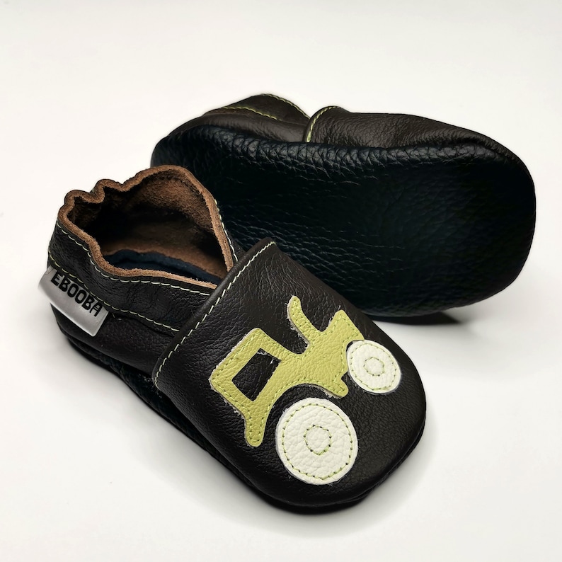 May include: A pair of brown leather baby shoes with a green tractor design. The shoes have a soft sole and are perfect for crawling and walking.