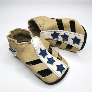 May include: A pair of tan leather baby sandals with blue star accents and a white strap. The sandals have an open toe and a velcro closure. The brand name "Ebooba" is visible on the strap.