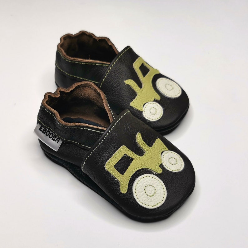 May include: A pair of black leather baby shoes with a green tractor design. The shoes have a velcro strap and the brand name "Ebooba" is printed on the side.