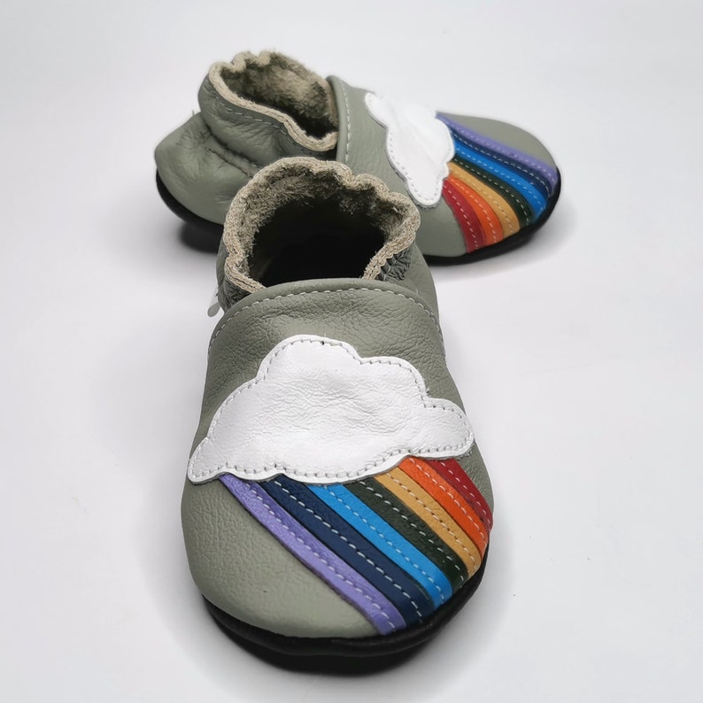 May include: A pair of gray leather baby shoes with a white cloud and rainbow design. The shoes have a soft, flexible sole and are perfect for little feet.