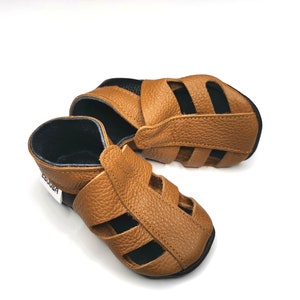 May include: A pair of brown leather sandals with black straps and a cut-out design. The sandals have a label that says "ABRODA".