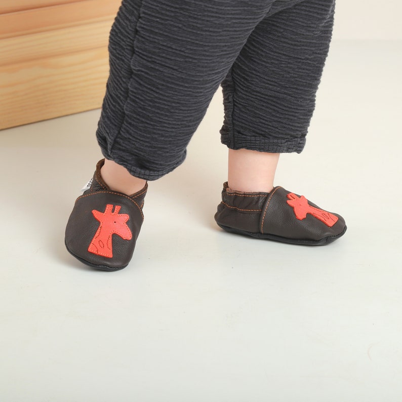 May include: Brown leather baby shoes with a red giraffe design. The shoes are soft and flexible, perfect for little feet.