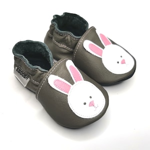 May include: A pair of gray leather baby shoes with a white bunny face and pink ears. The shoes have a green lining and the word "Ebooba" is printed on the side.