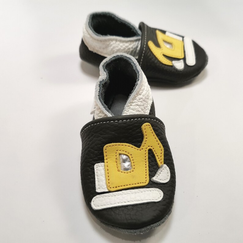 May include: Black leather baby shoes with a yellow and white excavator design. The shoes have a white lining and a gray sole.