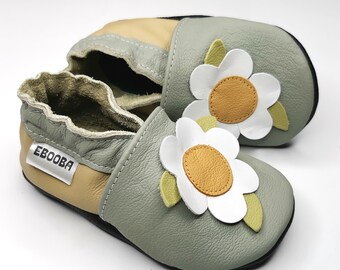 Può includere: Un paio di scarpine per neonati grigie e beige con un design a fiori bianchi. Le scarpine hanno una suola morbida e flessibile e sono realizzate in pelle. Il nome del marchio "Ebooba" è stampato sul lato della scarpa.