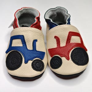 Peut inclure: Une paire de chaussons pour bébé avec un extérieur crème. L'une des chaussures présente un motif de tracteur bleu, l'autre un tracteur rouge. Les chaussures ont des roues noires et des accents rouges et bleus. L'intérieur est gris.