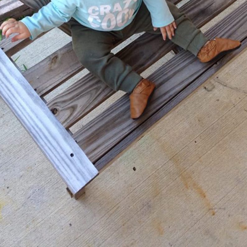 May include: A baby wearing a light blue shirt with the text "CRAZY COOL" in white letters, green trousers, and brown leather baby shoes. The baby is sitting on a wooden platform.