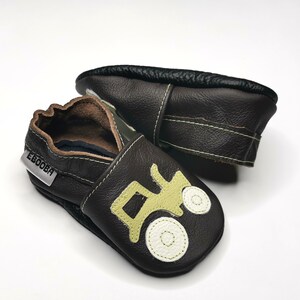 May include: Brown leather baby shoes with a green tractor design. The shoes have a soft sole and a velcro closure. The brand name "EEBOOBA" is printed on the side of the shoe.