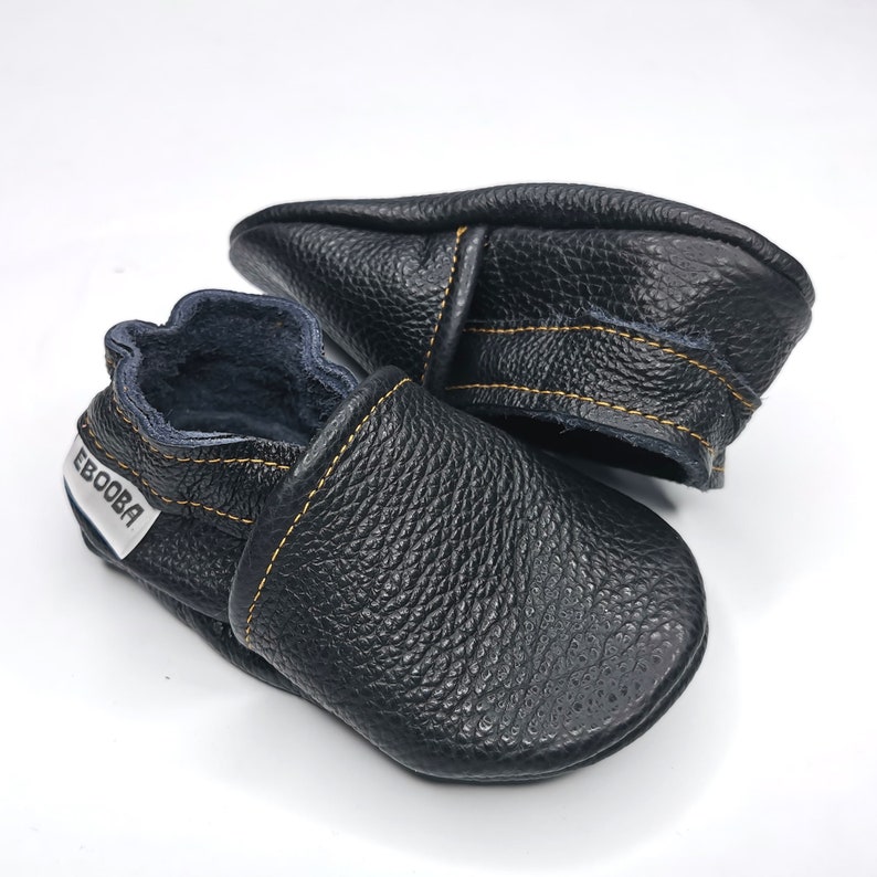 May include: A pair of black leather baby shoes with yellow stitching and a white label that says "Ebooba".