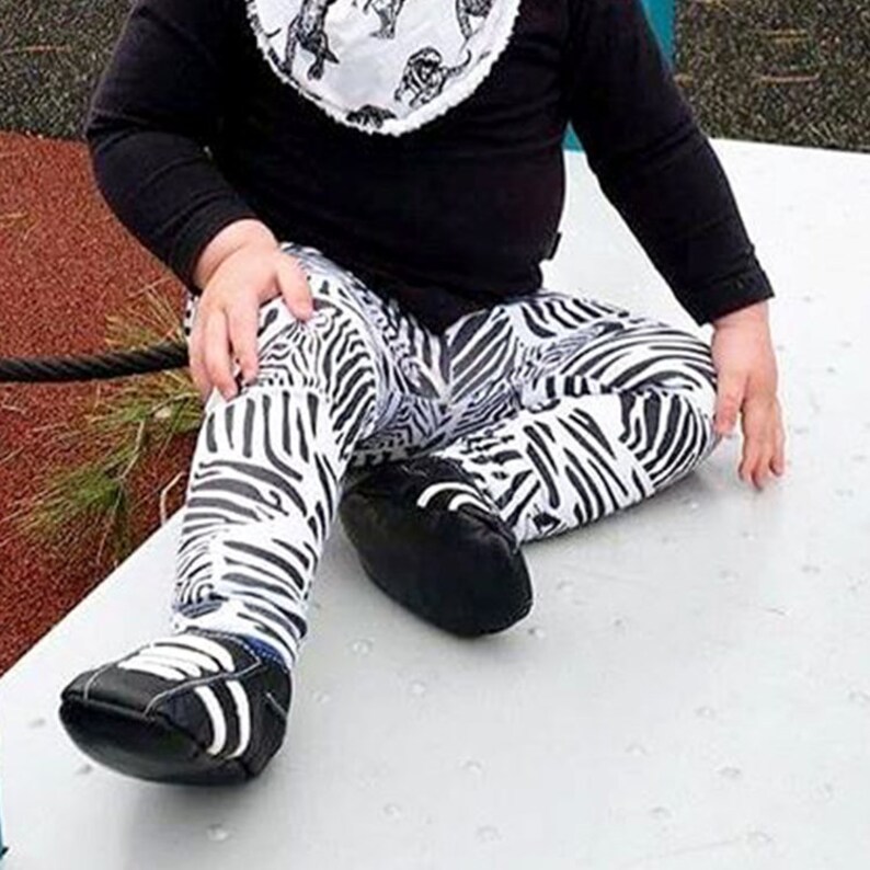 May include: A baby wearing black and white zebra print leggings and black shoes. The leggings have a wide, horizontal stripe pattern.