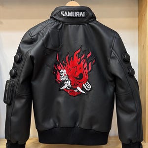 Könnte beinhalten: Eine schwarze Jacke in Lederoptik mit einer großen rot-weißen Stickerei einer stilisierten Maske auf dem Rücken. Der Kragen hat einen Aufnäher mit dem Text SAMURAI. Die Ärmel haben schwarze runde Akzente und eine kleine Reißverschlusstasche.