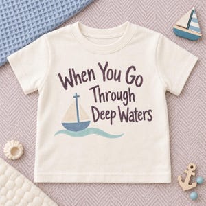 May include: A cream-colored short-sleeve t-shirt featuring the dark brown text When You Go Through Deep Waters, accompanied by a graphic of a blue and beige sailboat floating on a light blue wavy line, centered on the front of the garment.