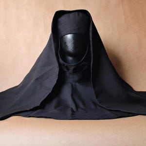 Può includere: Un copricapo nero a tinta unita caratterizzato da un cappuccio strutturato e un velo drappeggiato che si estende sulle spalle. L'indumento è esposto su una testa di manichino scura, mostrando una consistenza del tessuto liscia e semplice.