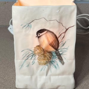 May include: A white ceramic luminary lamp with a textured, paper-bag-like shape. The front features a painted illustration of a small brown and black bird perched on a pine branch with two pinecones. The lamp emits a warm glow from within.