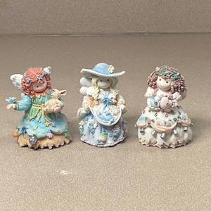 May include: Three decorative figurines featuring stylized characters. The left figure has red hair and a blue dress with stars. The center figure wears a blue hat and a patterned dress. The right figure has brown hair and a white dress with floral accents.