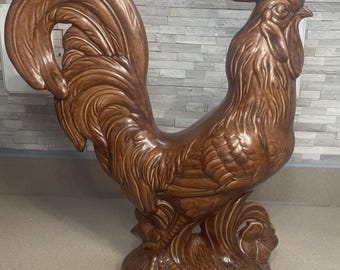 May include: A brown ceramic rooster figurine standing on a textured base. The sculpture features detailed molded feathers on the body and tail, a prominent comb on the head, and a small flower accent at the base. The finish is a glossy, solid brown.