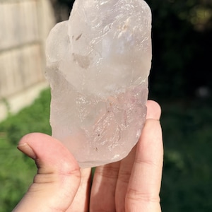 Pode incluir: Um cristal de quartzo rosa bruto e translúcido com um tom rosa pálido e uma textura de superfície irregular e irregular. A pedra apresenta fraturas e inclusões internas naturais, exibindo uma aparência turva e semitransparente.