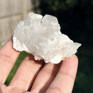 Pode incluir: Um aglomerado de cristais de quartzo branco translúcido com uma formação irregular e serrilhada. O espécime mineral apresenta múltiplas terminações pontiagudas e uma base branca opaca e rugosa, exibindo estruturas cristalinas naturais.