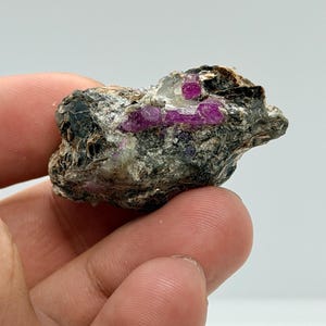 Peut inclure: Un spécimen minéral brut présentant des inclusions de cristaux magenta vif incrustées dans une matrice rocheuse sombre, texturée et marbrée de gris et de noir, avec des facettes cristallines visibles et des surfaces rugueuses.
