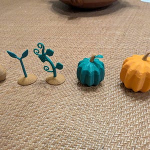 May include: A set of five small figurines depicting a plant life cycle, including a tan seed, a green sprout, a green vine, a teal pumpkin, and an orange pumpkin. Each piece is crafted with a textured finish and stands on a small base.