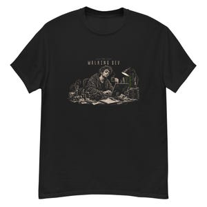 Può includere: Una t-shirt nera con un'illustrazione grafica di una persona che lavora a una scrivania con un portatile, una lampada e una tazza. Il testo sopra l'illustrazione recita THE WALKING DEV. Il design è realizzato in tonalità di grigio, nero e bianco.