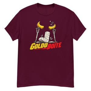 Puede incluir: Una camiseta de manga corta color granate con un gráfico de una escayola médica blanca en un pie, dos muletas y dos cuernos amarillos. Sobre la escayola hay pequeñas estrellas y un remolino. El texto "GOLDORAK BOÎTE" aparece en letras amarillas y rojas.