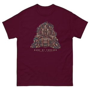 Peut inclure: Un t-shirt à manches courtes bordeaux arborant un graphisme représentant un trône composé de câbles informatiques et de composants matériels. Le motif inclut le texte "GAME OF THREADS" en lettres capitales blanches sous le trône illustré.