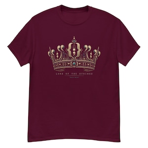 Op de afbeelding: Een bordeauxrood t-shirt met korte mouwen en een goudkleurige kroonafbeelding bestaande uit programmeersyntax-symbolen zoals accolades en puntkomma's. Onder de kroon staat de tekst LORD OF THE STRINGS en Hello World.