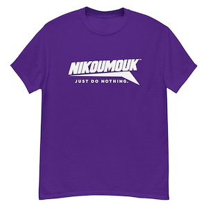 Puede incluir: Una camiseta morada de manga corta con un estampado gráfico blanco en el pecho. El diseño incluye el texto NIKOUMOUK en una fuente negrita e inclinada sobre la frase JUST DO NOTHING, junto a un elemento gráfico blanco afilado y angular.