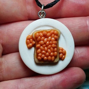 May include: A quirky pendant depicting a miniature plate with a slice of toast topped with baked beans. The pendant has a white plate, a black cord, and a silver-tone clasp. The beans are a reddish-brown colour.