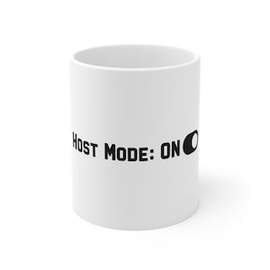 Może przedstawiać: Biały ceramiczny kubek z czarnym napisem "HOST MODE: ON" oraz ikoną przełącznika. Wzór jest nadrukowany poziomo na środku kubka, który ma gładki, cylindryczny kształt oraz prosty, przejrzysty wygląd.