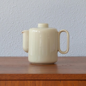 May include: A cream-coloured ceramic teapot with a cylindrical body, a short spout, a flat lid with a small knob handle, and a large, rectangular-shaped handle on the side. The teapot has a smooth, glossy finish and a simple, uniform design.
