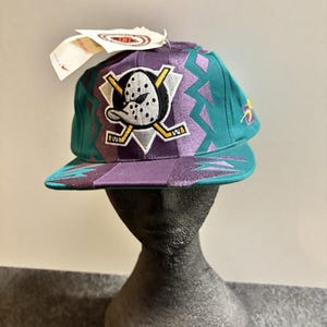 May include: A teal and purple baseball cap featuring an embroidered Anaheim Ducks hockey logo on the front. The cap has a flat brim, a geometric pattern, and a white tag attached to the top. The design includes two crossed yellow hockey sticks.