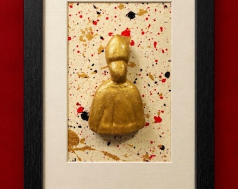 Op de afbeelding: Een ingelijst wandkunstwerk met een driedimensionaal goudkleurig figuur van een persoon met een hoge hoed, gecentreerd op een crèmekleurig canvas met verspreide rode, zwarte en gouden verfspatten, in een zwarte lijst.