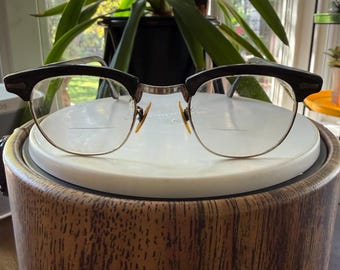Puede incluir: Gafas de estilo browline con montura superior negra y bordes inferiores finos de tono plateado. Las lentes incluyen una línea bifocal horizontal visible. El puente y las plaquetas son metálicos, con un acabado en tono ámbar.