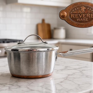 Puede incluir: Un cazo de acero inoxidable con base de cobre y tapa de cristal transparente. El mango presenta una pieza de acento de color cobre. Una placa con el logotipo ovalado muestra el texto REVERE WARE con una ilustración de perfil.