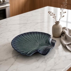Puede incluir: Un plato de servicio de cerámica con forma de concha marina grande con un acabado brillante en tonos azul profundo y verde azulado. El plato presenta crestas prominentes y radiantes que imitan la textura natural de una vieira.