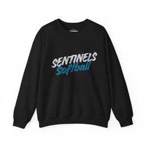 May include: A black long-sleeve crewneck sweatshirt featuring the text SENTINELS in white and Softball in blue across the chest. The lettering has a distressed, brush-stroke texture. The garment has a ribbed collar, cuffs, and hem.